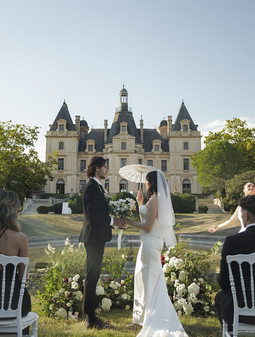 Cérémonie de mariage en extérieur devant un château avec ombrelle personnalisée.