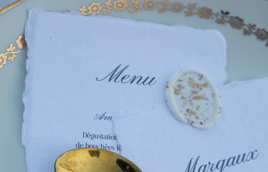 Menu de mariage en papier artisanal avec sceau de cire doré, posé sur vaisselle ancienne.
